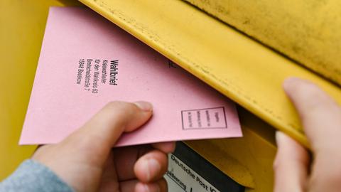 Eine Hand wirft einen rosa Briefwahlumschlag in den Schlitz eines gelben Briefkastens.