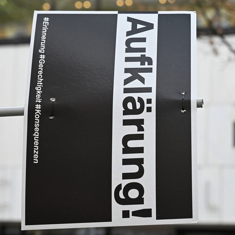 Im Bildvordergrund ein Schild mit dem Schriftzug "Aufklärung" - auf der Seite liegend. Im Hintergrund unscharf das Gebäude des Landtags.
