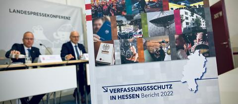 Broschüre Verfassungsschutzin Hessen - Bericht 2022 - im Hintergrund sitzt Innenminister Beuth - an der Wand steht Landespresskonferenz