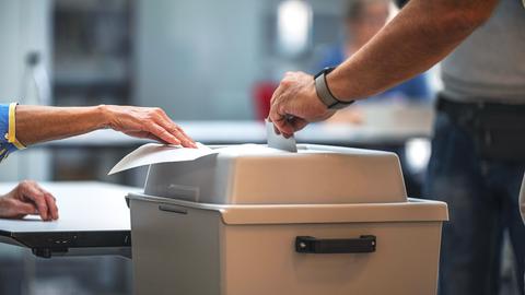 Eine Hand steckt einen Umschlag in eine versiegelte Wahlurne