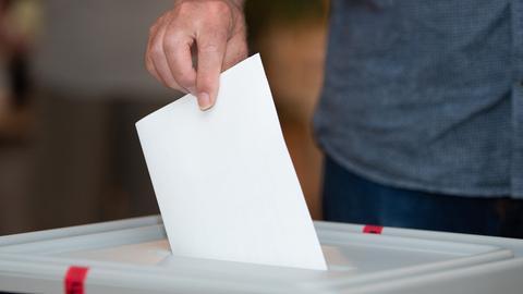 Eine Hand eines Wählers wirft einen Stimmzettel in die Wahlurne.