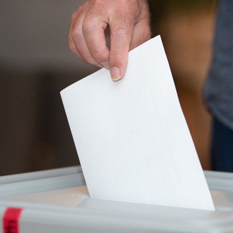 Eine Hand eines Wählers wirft einen Stimmzettel in die Wahlurne.