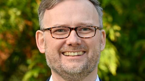 Jens Womelsdorf: Mann mit dunkler Anzugjacke, Brille, kurzen, grau-dunkelblonden Haaren und einem kurzen Bart