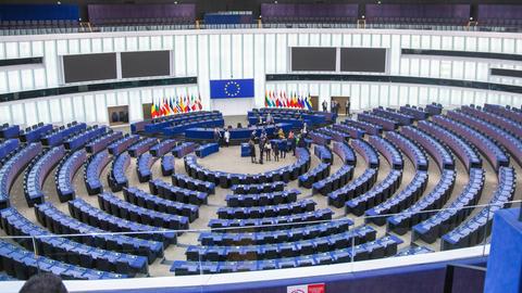 Foto eines großen Plenarsaals mit vielen blauen Sitzen und einer EU-Flagge hinter dem vorderen Pult. In der Mitte vor dem Stehpult für Vortragende stehen einige Menschen locker beisammen.