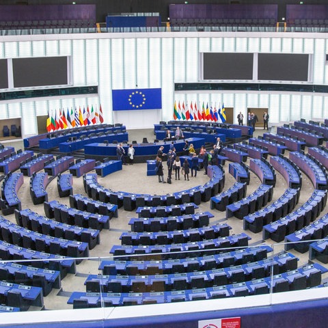 Foto eines großen Plenarsaals mit vielen blauen Sitzen und einer EU-Flagge hinter dem vorderen Pult. In der Mitte vor dem Stehpult für Vortragende stehen einige Menschen locker beisammen.