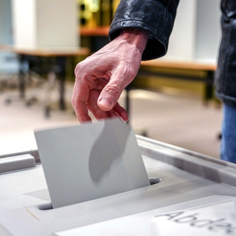 Eine Hand steckt einen Stimmzettel in eine Wahlurne. Die Person trägt eine schwarze Jacke. Im Hintergrund sind Tische und Wahlkabinen zu sehen. Ein Teil des Texts auf der Wahlurne ist sichtbar: „Abde...“.