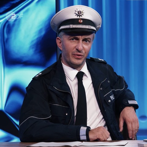 Moderator Böhmermann mit Polizei-Mütze an Moderationstisch