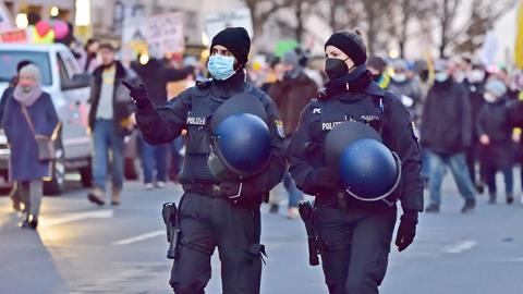 Im Bildvorgrund zwei Polizeibeamte mit Helmen in der Hand. Im Hintergrund unscharf eine Demonstration.