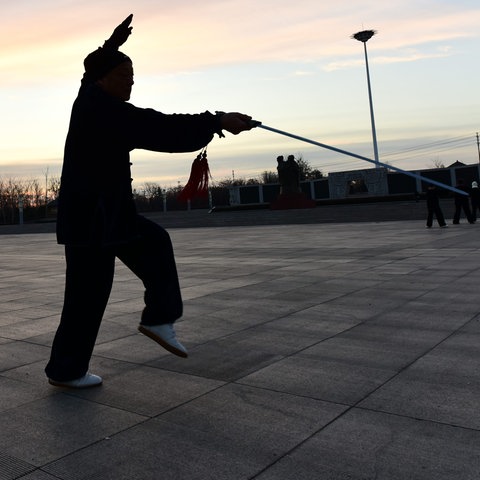 Eine schwarz gekleidete Person bewegt sich auf einem Platz mit einem Schwert. Das Bild wurde gegen die untergehende Sonne fotografiert, so dass es als Schattenriss erscheint.