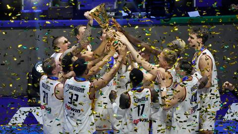 Basketballspieler in weißen Trikots halten einen goldenen Pokal hoch, um sie herum wirbelt goldenes Konfetti.