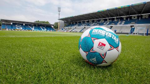 Ein Fußball liegt im Vordergrund des Bildes auf dem grünen Rasen des Stadions von Darmstadt 98. Im Hintergund leere Tribünen auf denen "SVD" zu lesen ist.