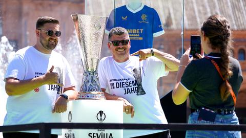Zwei Männer mit Sonnenbrillen stehen neben einem großen Pokal, zeigen auf ihn und lachen in die Kamera.