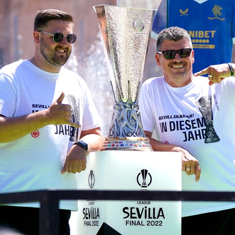 Zwei Männer mit Sonnenbrillen stehen neben einem großen Pokal, zeigen auf ihn und lachen in die Kamera.