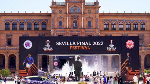 Vor einem historischen Gebäude ist eine große Bühne mit der Aufschrift "Sevilla Final 2022 Festival" aufgebaut. Davor Menschen, Autos und Blumen.