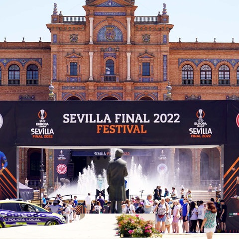 Vor einem historischen Gebäude ist eine große Bühne mit der Aufschrift "Sevilla Final 2022 Festival" aufgebaut. Davor Menschen, Autos und Blumen.