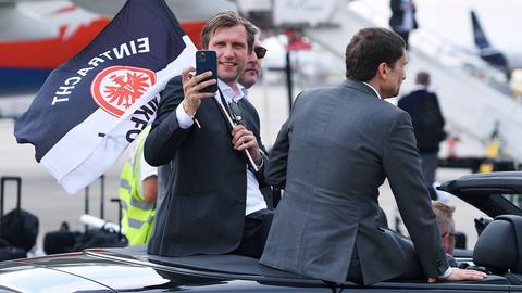Markus Krösche sitzt mit zwei weiteren Menschen erhöht in einem Cabrio, hält eine Eintrachtfahne und ein Mobiltelefon in der Hand -  in Richtung Kamera, als würde er die Fotografierenden fotografieren.