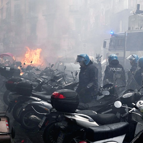 In einer Straße sind viele Zweiräder, viele Polizisten mit Helmen, ein großer Einsatzwagen der Polizei im Bildvordergrund scharf zu sehen. Im Bildhintergrund unscharf ein brennendes Auto und viele Rauchschwaden und Nebel überall.