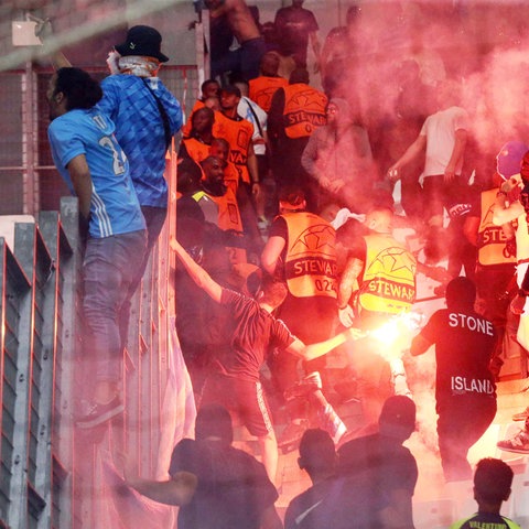 Menschen auf der Tribune eines Stadions in Aufruhr. Inmitten eine Leuchtrakete, die die Szenerie beleuchtet.