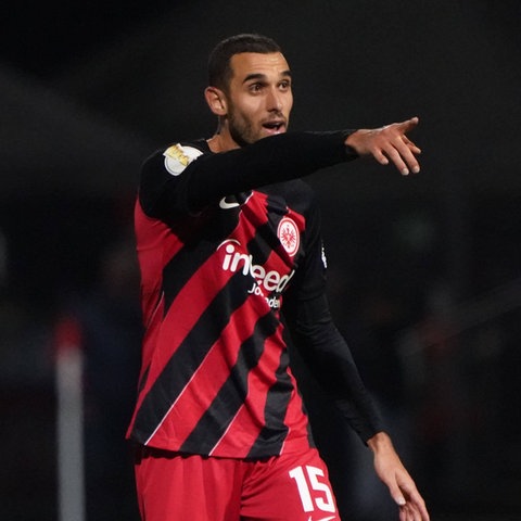 Ein Mann im schwarz-rot gestreiften Trikot zeigt - auf dem Spielfeld stehend - mit dem Arm in eine Richtung.