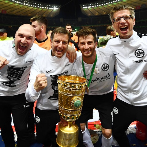 Vier Männer stehen um einen goldenen Pokal in einem Stadion und jubeln.
