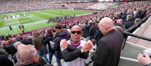 Im Bildvordergrund ein West Ham United Fan, der seine Arme vor seinem Oberkörper überkreuzt und in die Kamera schaut. Im Bildhintergrund ein volles Fußballstadion.