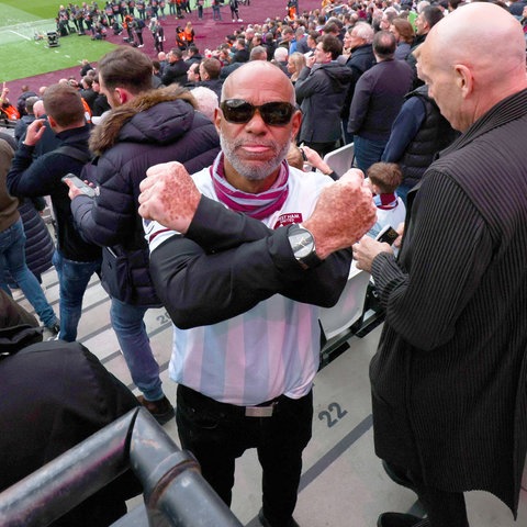 Im Bildvordergrund ein West Ham United Fan, der seine Arme vor seinem Oberkörper überkreuzt und in die Kamera schaut. Im Bildhintergrund ein volles Fußballstadion.