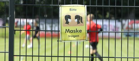 Ein provisorisches Schild mit der Aufschrift "Bitte Maske tragen" hängt an einem Zaun eines Amateurfußballfeldes.