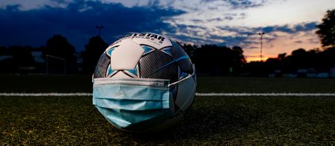 Ein Fußball mit Mundschutz liegt in der Abenddämmerung auf einem Spielfeld.