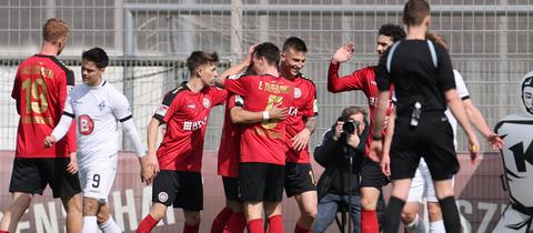 Mehrere Spieler vom SV Wehen Wiesbaden (rote Trikots) jubeln gemeinsam über ein Tor. 