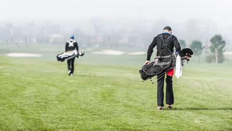 Zwei Männer, die ihre Golftaschen über den Platz tragen, von hinten.