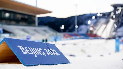 Im Vordergrud: Ein Banner auf dem "Beijing 2022" steht. Im Hintergrund verschwommen: eine Wintersportanlage in Peking.