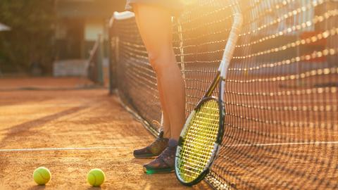 Auf einem Tennis-Platz liegen Bälle. Ein Schläger lehnt am Netz. Die Beine einer jungen Tennisspielerin sind zu sehen.