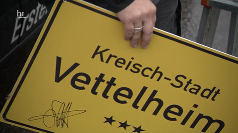 Eine Frau hält ein Ortsschild mit dem Namen "Kreischstadt Vettelheim".