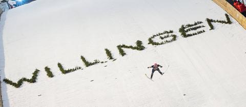 Ein Skispringer fliegend vor einer weißen Schneepiste, auf welcher in grünen Buchstaben "Willingen" steht. 