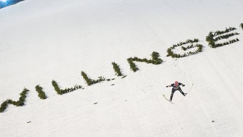 Ein Skispringer fliegend vor einer weißen Schneepiste, auf welcher in grünen Buchstaben "Willingen" steht. 