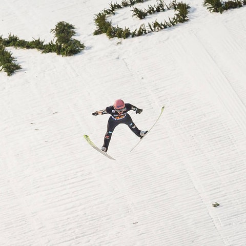 Ein Skispringer fliegend vor einer weißen Schneepiste, auf welcher in grünen Buchstaben "Willingen" steht. 