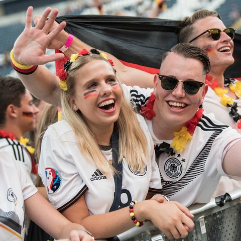 Zwei Menschen schauen in die Kamera, Deutschlandfahnen in der Hand, schwarz-rot-gold geschminkt. Sie stehen in einer Menschentraube, die beim Public Viewing einem Fußballspiel zuschaut.