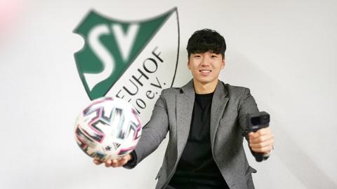 Jeong-Jin Lee steht mit einem Ball in der Hand vor einer Wand, an welcher das Signet des "SV Neuhof 1910 e.V." angebracht ist.