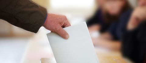 Hand mit einem gefalteten Wahlzettel, wirft den Wahlzetteel in eine Box