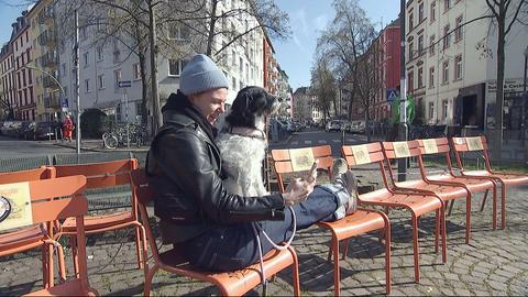 Eine Passantin genießt den Sonnenschein mit ihrem Hund auf einer Bank.