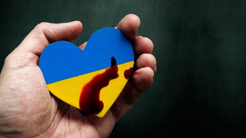 Eine Hand hält ein hölzernes Herz in den Nationalfarben der Ukraine, mit Blut beträufelt