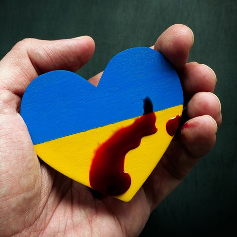 Eine Hand hält ein hölzernes Herz in den Nationalfarben der Ukraine, mit Blut beträufelt