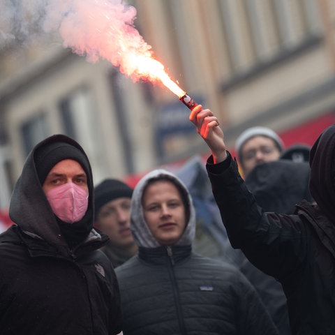 Dunkel gekleidete Menschen in einer Gruppe auf der Straße. Eine Person hält ein rauchendes Leuchtfeuer in der Hand.