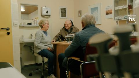 Ein Arzt sitzt mit zwei Patienten in seinem Behandlungszimmer am Schreibtisch.