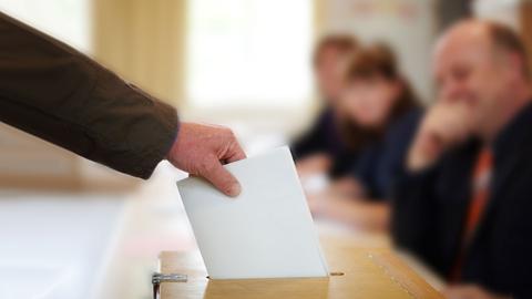 Eine männliche Hand steckt Wahlunterlagen in eine Wahlurne