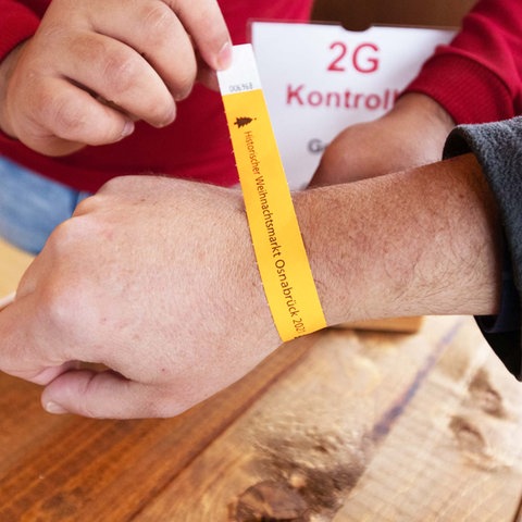 Zwei Hände legen einer dritten Hand ein orangfarbenes Papierbändchen um das Handgelenk mit der Aufschrift "Weihnachtsmarkt Osnabrück".