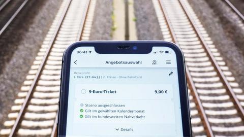 Das Bild zeigt ein Smartphone im Bildvordergrund und Bahngleise unscharf im Bildhintergrund. Auf dem Display ist eine Nahverkehrsapp mit Anwahl des Buttons "9-Euro-Ticket" zu sehen. 