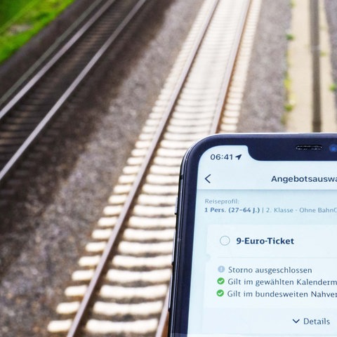 Foto menunjukkan smartphone di latar depan dan rel kereta api tidak fokus di latar belakang.  Di layar aplikasi transfer lokal dengan tombol yang dipilih "tiket 9 euro" untuk melihat. 