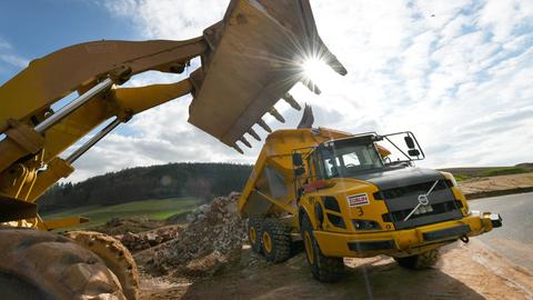 Eine Baggerschaufel ragt von links ins Bild. Durch deren Zinken bricht die strahlende Sonne. Ein LKW lädt Schutt ab (mittig im Bild). Er steht neben einer noch unfertigen Straße. 