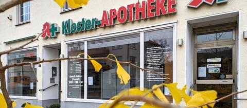 Außenansicht einer Apotheke. Schaufenster und Schriftzug "Kloster-Apotheke" mit Apothekenzeichen und Kreuz daneben. 